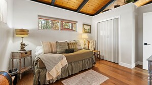 A bedroom with a wooden ceiling, a bed with a ruffled skirt, a patterned rug, and a window with a view of trees.