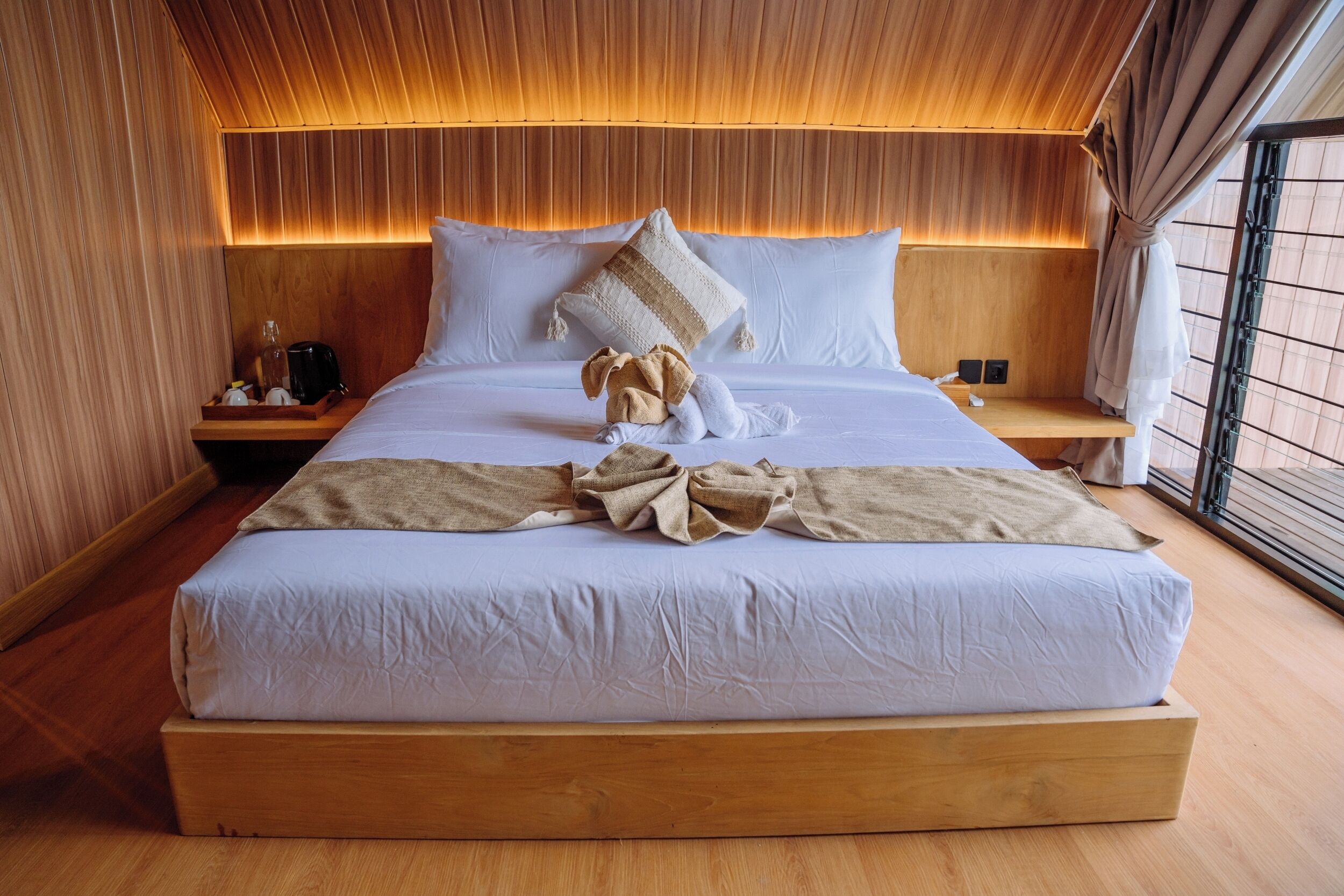 Een groot bed met witte lakens, een houten hoofdeinde en een nachtkastje met een lamp en een fles.