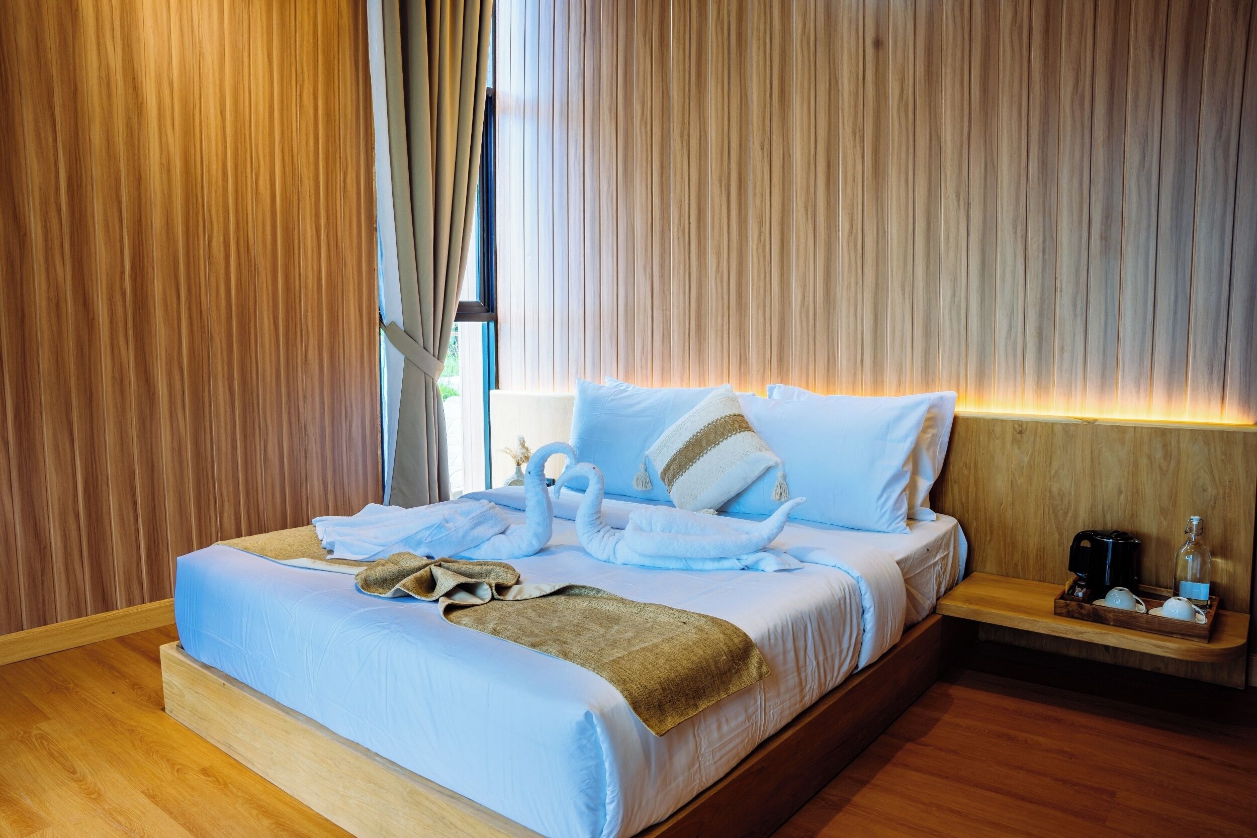 A neatly made bed with a blue bedspread and white pillows, a wooden bedside table with a coffee maker and a bottle, and a wooden floor.