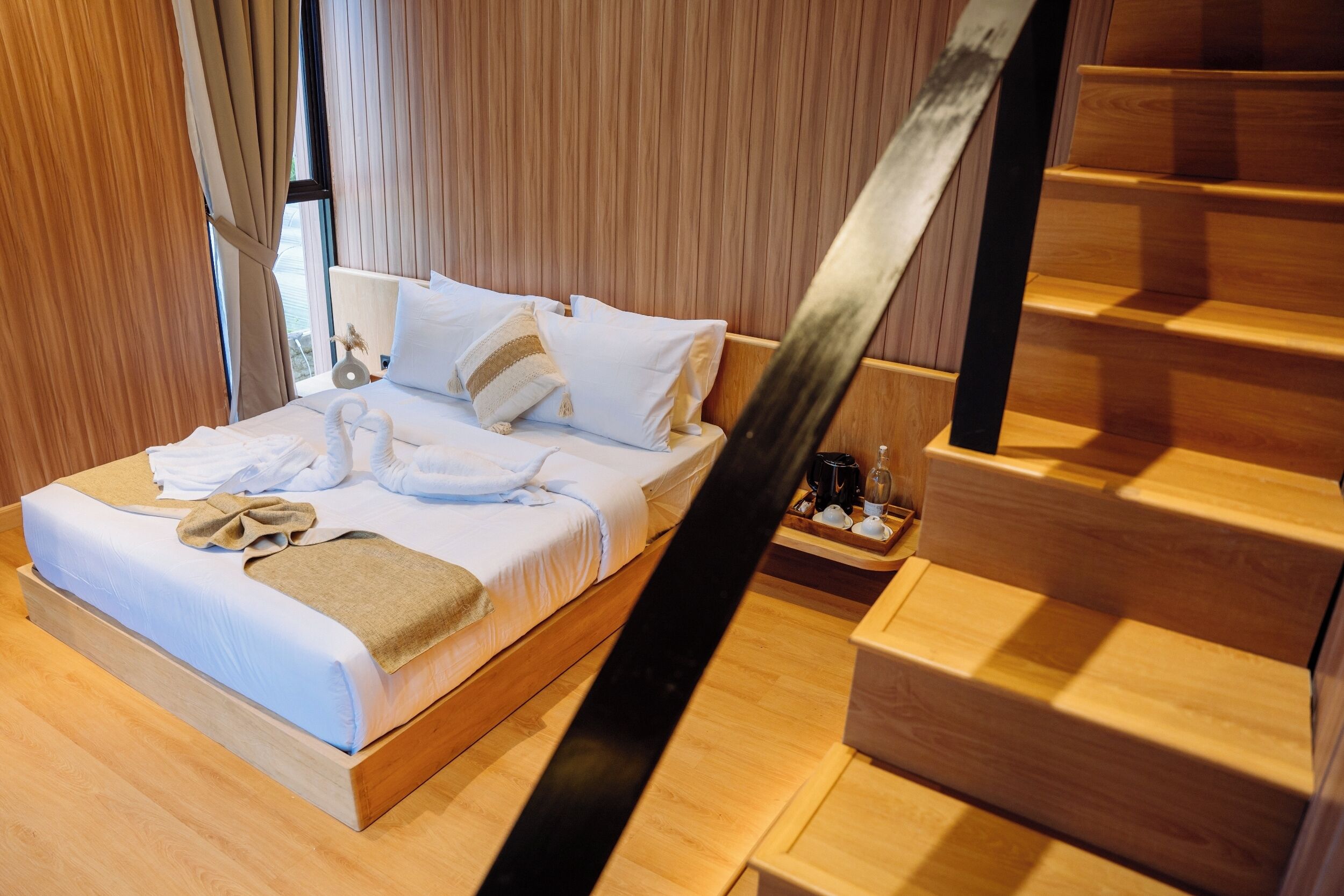 A neatly made bed with white linens and a brown throw blanket, a wooden nightstand with a glass of water and a cup, and a wooden staircase with a black handrail.