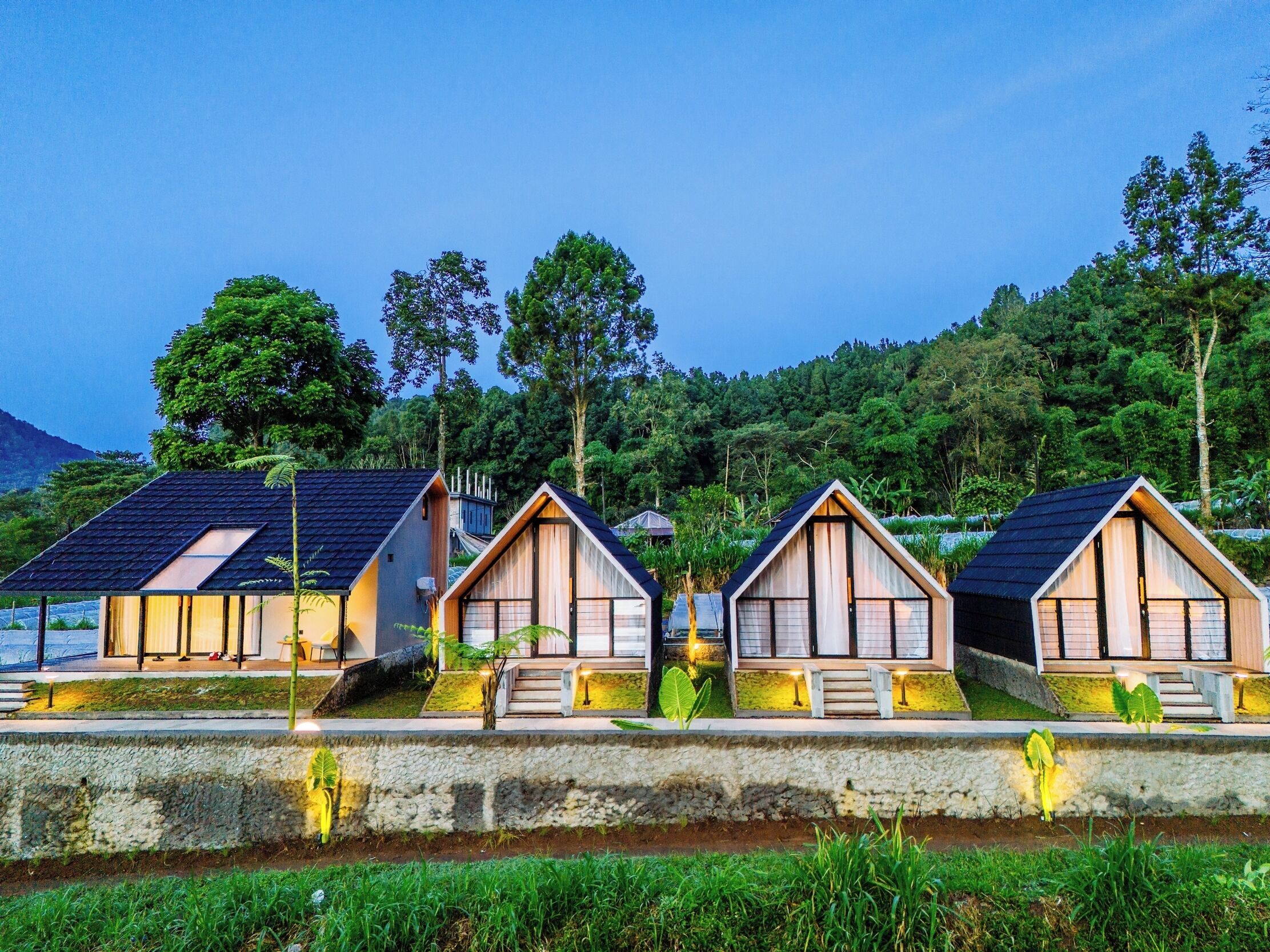 Una hilera de cabañas pequeñas y modernas con techos inclinados, ubicadas sobre una plataforma de concreto con vista a un bosque frondoso y una montaña al fondo.