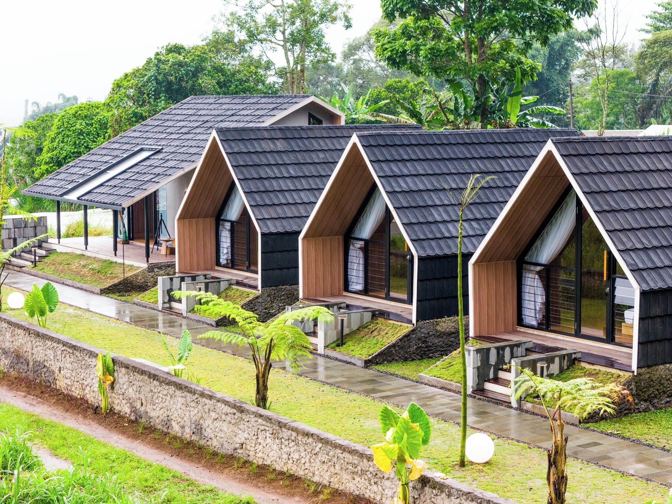 A row of modern, triangular-roofed cabins with wooden exteriors and glass doors, set in a lush, green environment with a stone pathway and small plants.