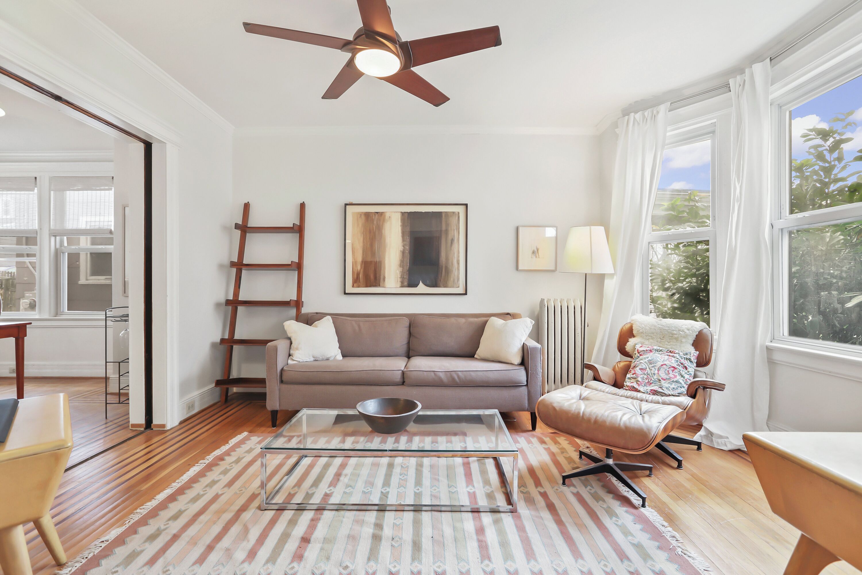 A modern living room with a glass coffee table, a brown leather sofa, a rocking chair, and a ceiling fan.