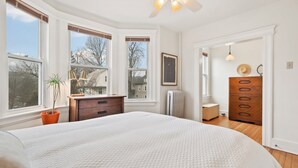 Een slaapkamer met een groot bed, een dressoir, een radiator en een raam met uitzicht op huizen.
