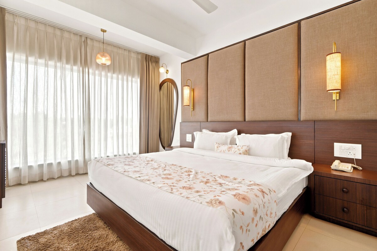 Een moderne hotelkamer met een groot bed, een houten hoofdbord, een nachtkastje met een lamp, en een groot raam met gordijnen.