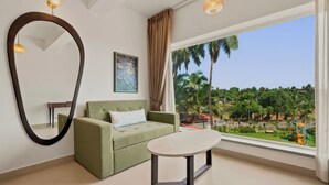 A modern room with a sofa, a table, a large mirror, and a window with a view of palm trees and a playground. - Verano Hotel & Spa  (Arpora)