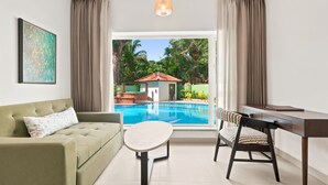 A room with a sofa, a dining table, a chair, a desk, a hanging light, and a view of a swimming pool and greenery outside. - Verano Hotel & Spa  (Arpora)