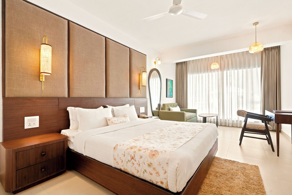 A hotel room with a large bed, bedside tables, a desk, a chair, a ceiling fan, and a large window with curtains.