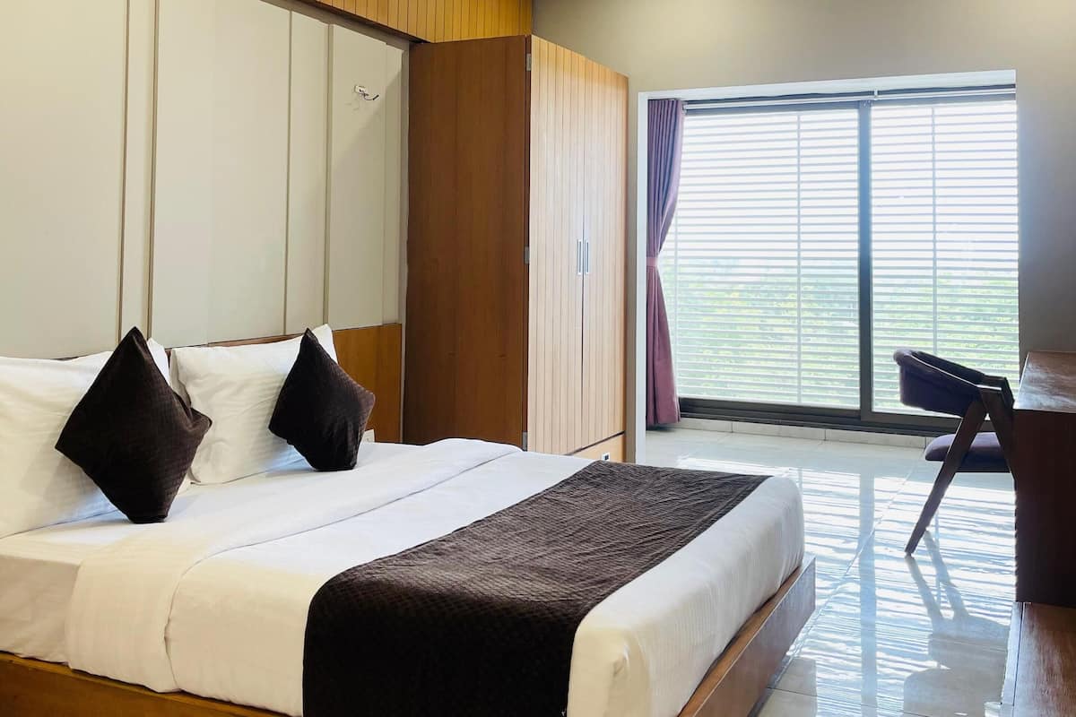 A modern hotel room with a large bed, wooden headboard, ceiling fan, and a view of the outdoors through large windows.