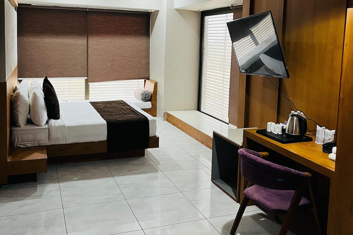 A modern hotel room with a bed, desk, chair, and a large window with blinds.