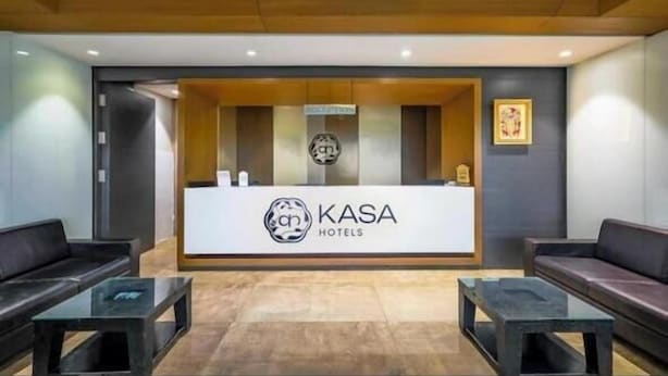 Un área de recepción con un mostrador identificado con el nombre "KASA HOTELS". Hay dos sofás negros y dos mesas con superficie de vidrio.