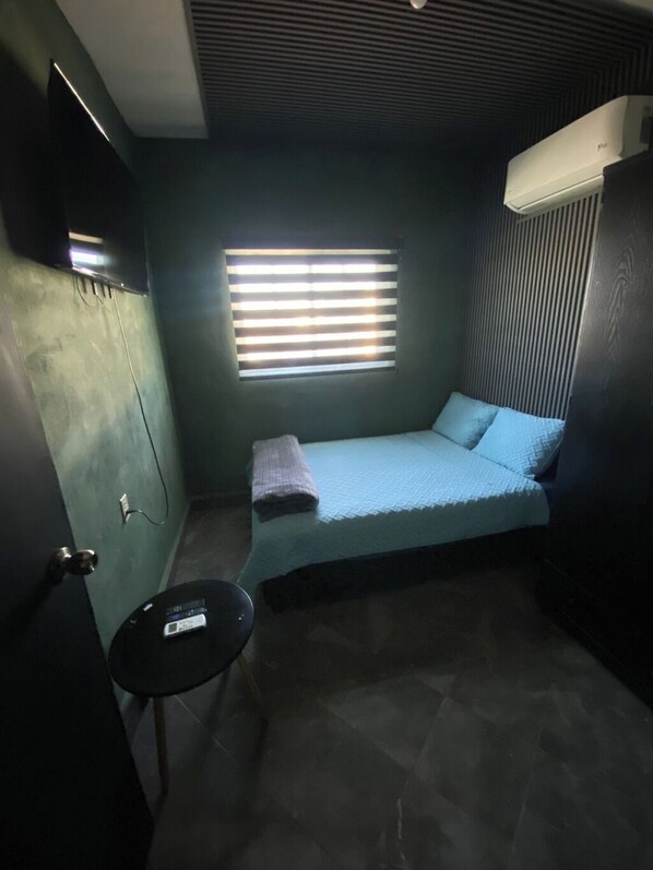 A small, dimly lit room with a single bed, a small table, and a window with blinds.