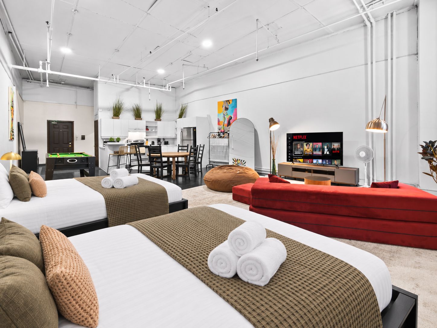 A modern hotel room with two beds, a red sofa, a dining area, and a TV displaying Netflix.