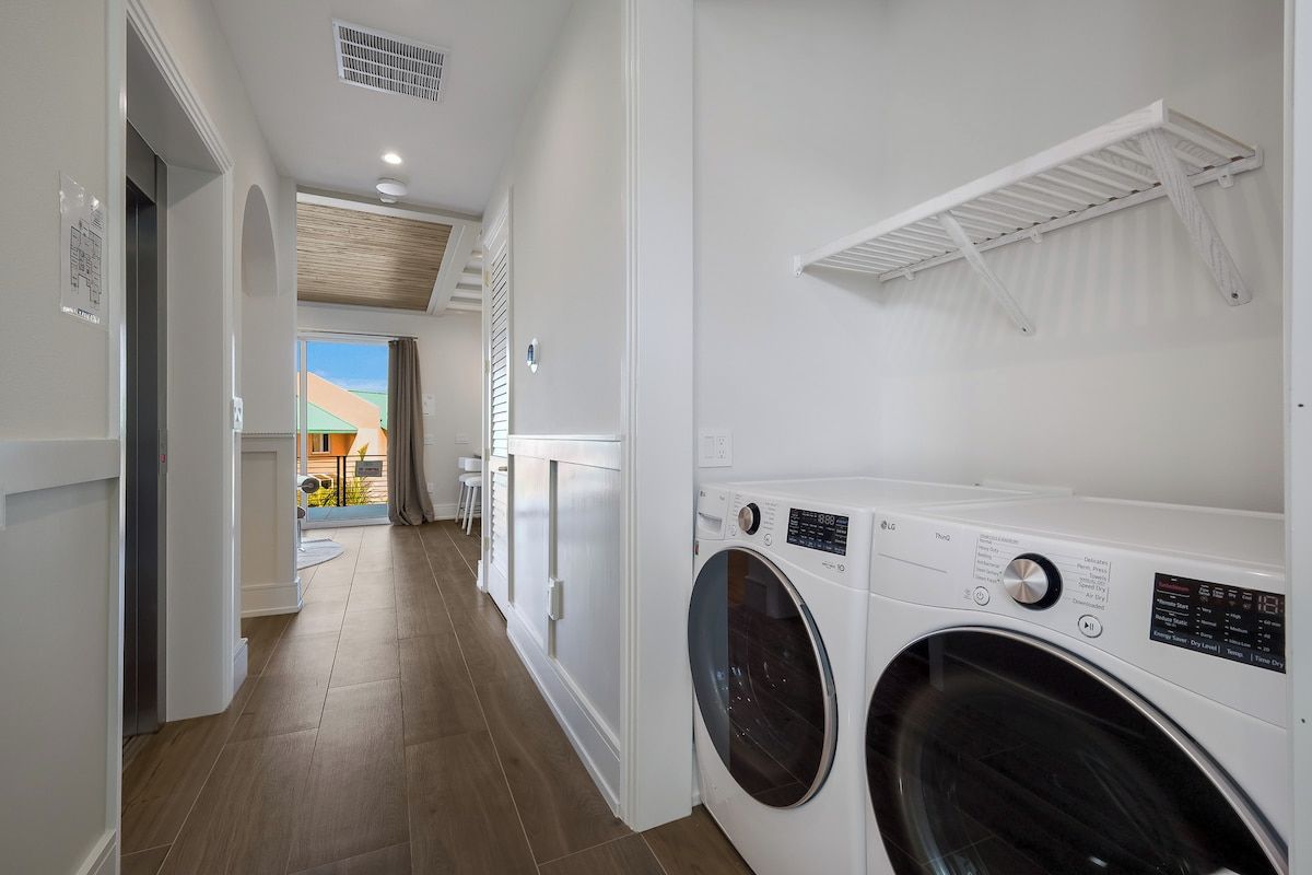 A laundry room with a washing machine and dryer, a white shelf, and a view of a house through an open door.