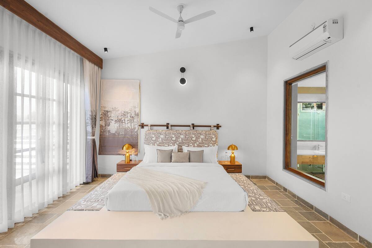Une chambre dotée d’un grand lit, d’un ventilateur de plafond, d’un climatiseur, d’une table de chevet avec une lampe et d’une fenêtre donnant sur une piscine.