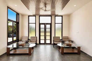 A modern waiting area with two glass-top tables, four patterned chairs, and a large window with a view of the outdoors. - Still River Suites with Heated Pool, Jacuzzi by ELIVAAS (Udaipur)
