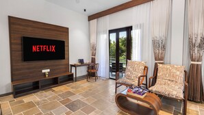 Una sala de estar con un televisor de pantalla plana mostrando 'NETFLIX', un mueble de madera para entretenimiento, un área de comedor con mesa y sillas, y un amplio ventanal corredizo que da a un espacio exterior.