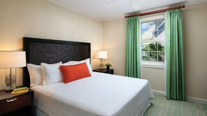 A neatly made bed with white bedding and a red accent pillow, flanked by two bedside tables with lamps and a small plant. The room features a large window with green curtains and a view of a house. - Harborside Resort at Atlantis, Bahamas 2 Bedrm Lockoff Villa  7/24/26 - 7/31/26 (Paradise Island)