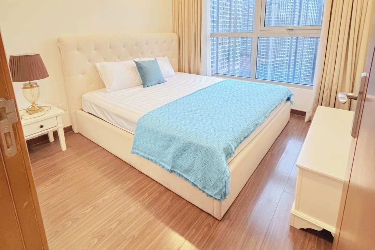 A hotel room with a large bed, a bedside table with a lamp, a wardrobe, and a window with a city view.