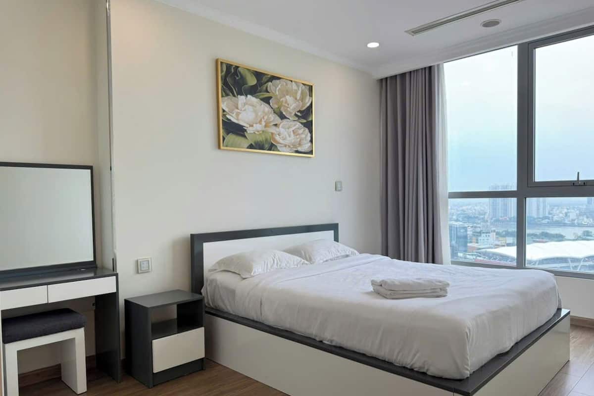 A modern bedroom with a large bed, a bedside table, a desk, and a window with a city view.