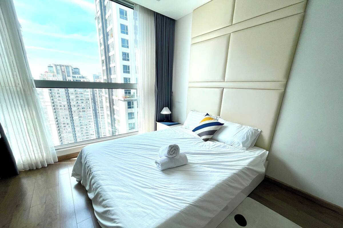 A hotel room with a large bed, a headboard, a nightstand, a lamp, and a view of tall buildings through the window.