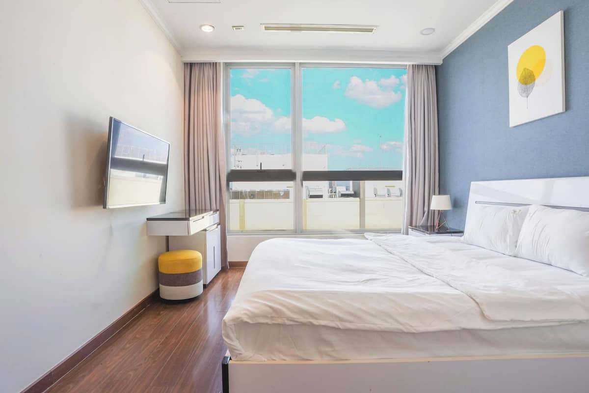 A hotel room with a large bed, a TV on the wall, a window with a view of the city, and a small table with a lamp.
