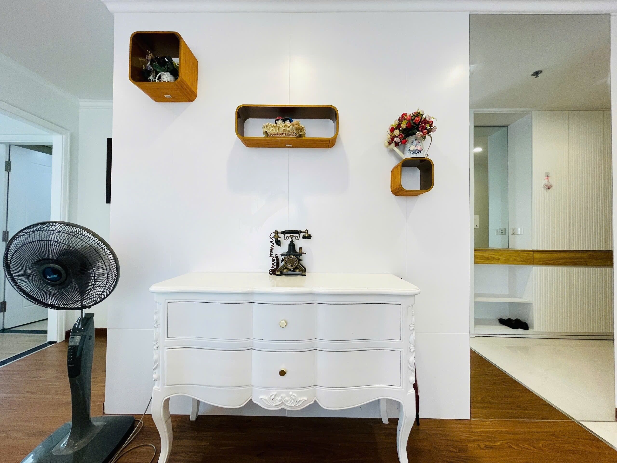 A white dresser with drawers, a vintage telephone, and wooden shelves with decorative items.