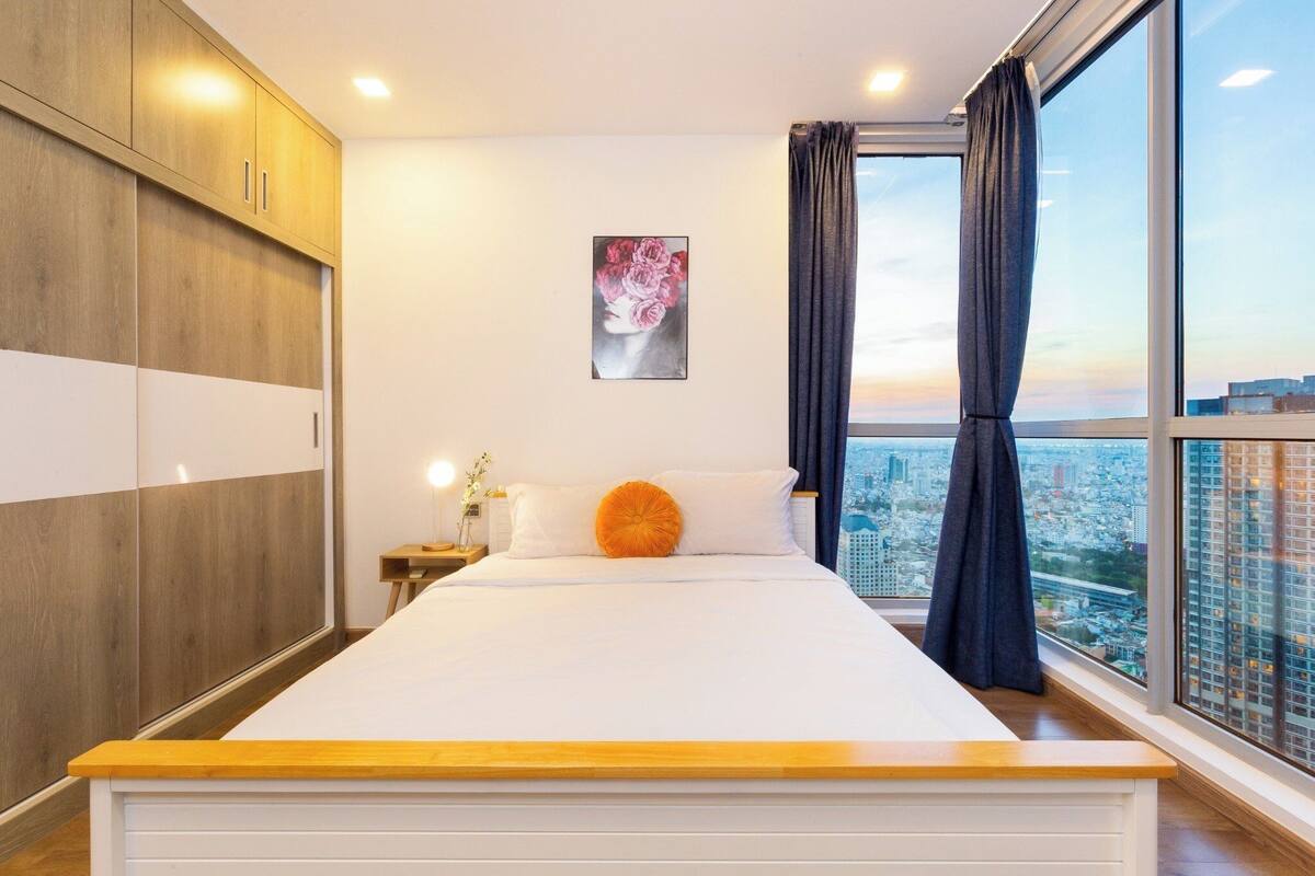 A bedroom with a large bed, a nightstand with a lamp, a painting on the wall, and a city view through the window.