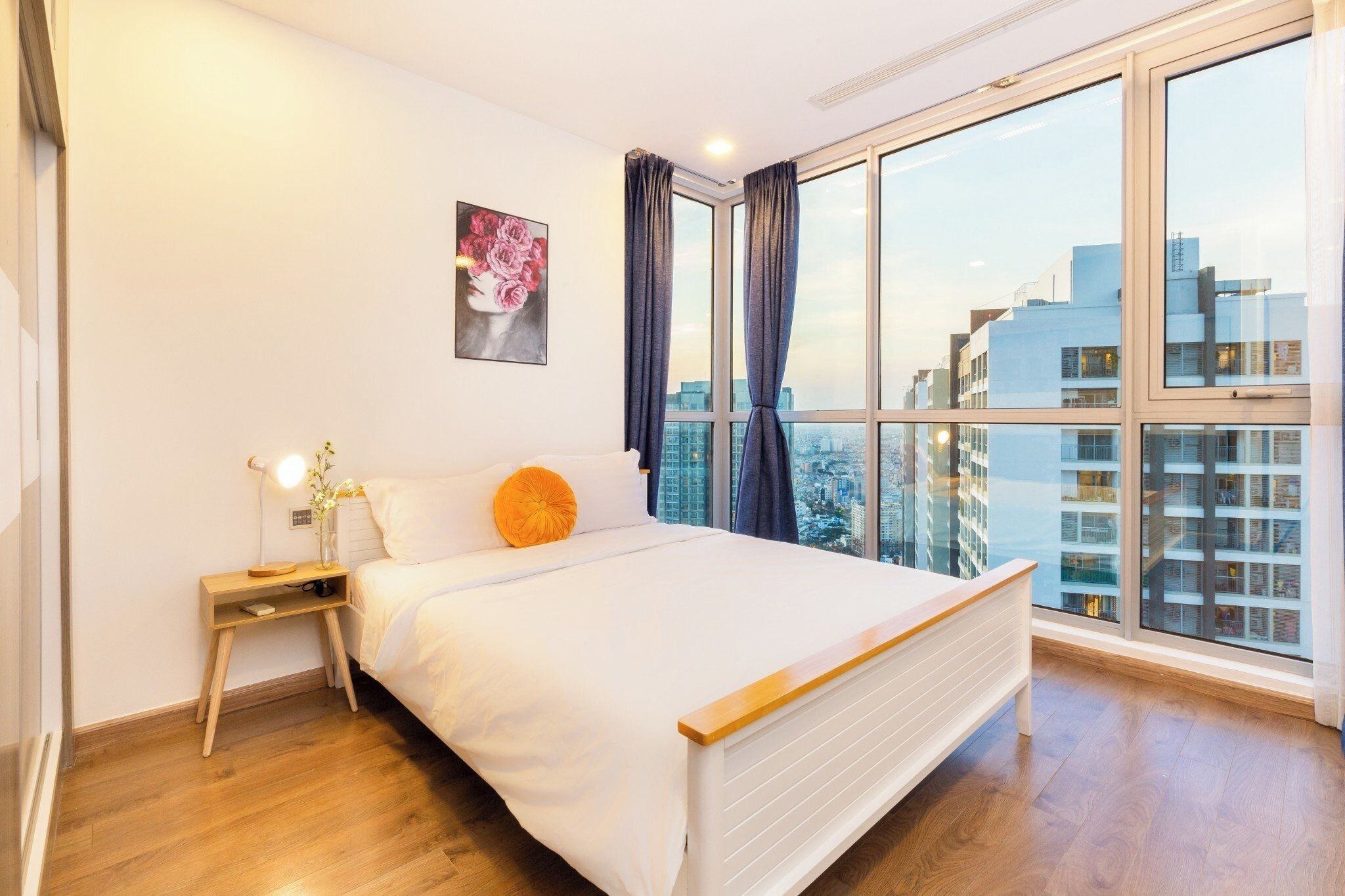A bedroom with a large bed, a bedside table with a lamp, a window with curtains, and a city view.