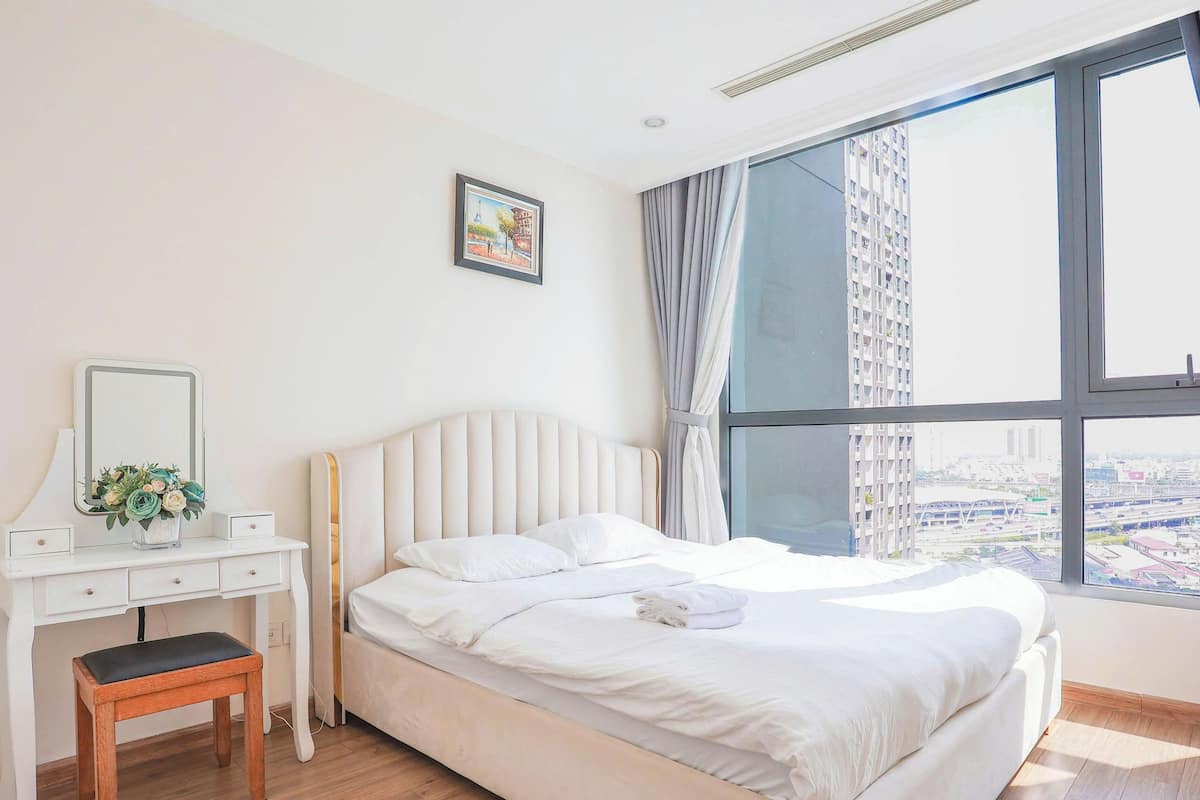 A bedroom with a bed, a vanity with a mirror, and a window with a city view.