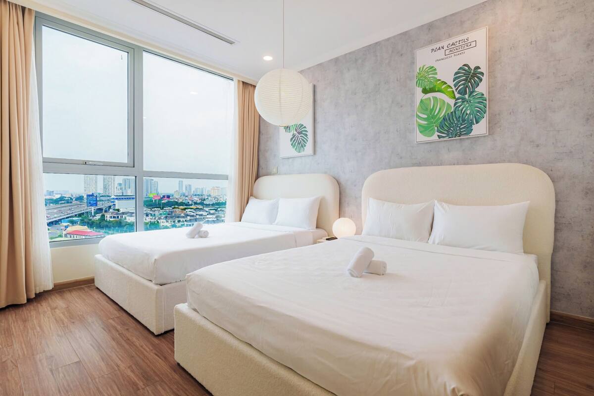 A hotel room with two beds, a large window with a city view, a plant wall art, and a hanging light fixture.