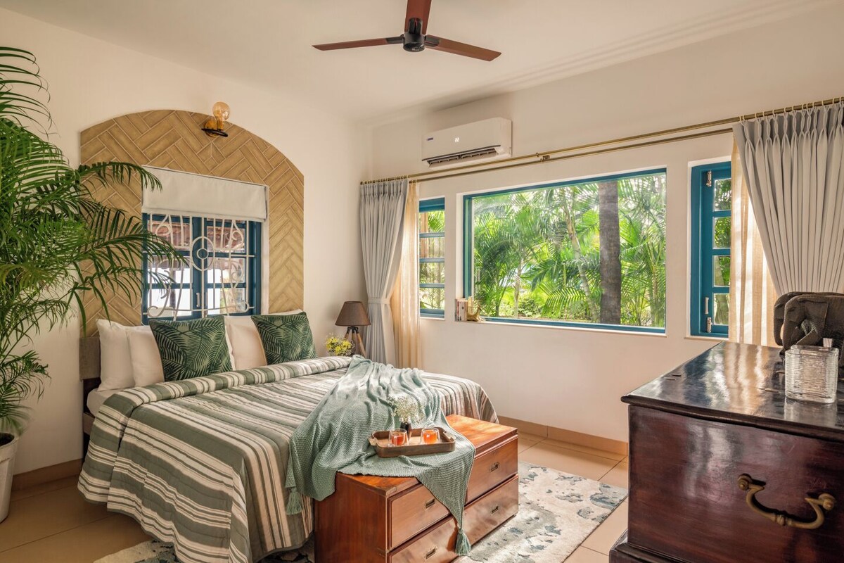 A bedroom with a bed, a dresser, a nightstand, a ceiling fan, and a window with a view of greenery.