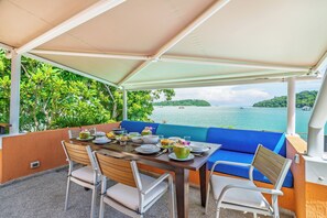 An outdoor dining area with a table set for two, surrounded by blue cushioned seating, overlooking a body of water with boats. - 5 BR Seaview Pool Villa Nambo in Panwa (Wichit)