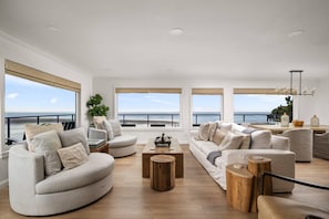 A spacious living room with a view of the ocean, featuring a large sofa, a coffee table, and a dining area. - Magnificent Oceanview Retreat w/ Hot Tub, Wraparound Deck & Luxe Primary Suite, Foosball, EV Charger (Tillamook)