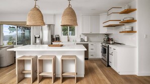 A modern kitchen with a central island, wooden stools, hanging woven light fixtures, white cabinetry, and wooden shelves.