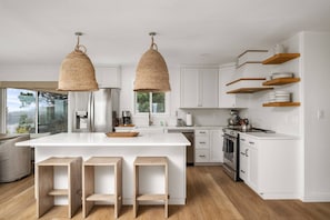 A modern kitchen with a central island, wooden stools, hanging woven light fixtures, white cabinetry, and wooden shelves. - Magnificent Oceanview Retreat w/ Hot Tub, Wraparound Deck & Luxe Primary Suite, Foosball, EV Charger (Tillamook)