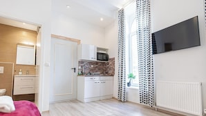 Moderni hotellihuone, jossa on pieni keittiönurkkaus, taulutelevisio ja suuri ikkuna, jossa on kuviolliset verhot.