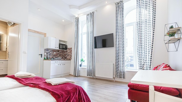Moderni hotellihuone, jossa on sänky, punainen sohva, keittiö sekä seinälle asennettu televisio.