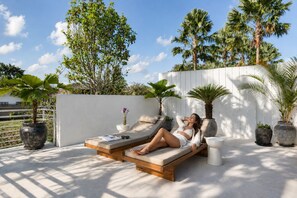A woman relaxing on a wooden lounge chair with a cushion, surrounded by potted plants and a white wall. - Modern Villa 2BR Casa Azuca by OriVista (Canggu)