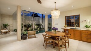 A modern kitchen with wooden cabinets, a dining area with a round table and chairs, a built-in island with a sink, and a large window with a view of the outdoors. - Modern Villa 2BR Casa Azuca by OriVista (Canggu)