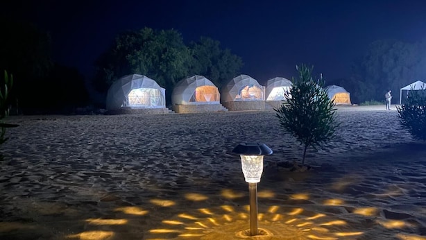 Une série de tentes en forme de dôme, illuminées la nuit, disposées en rangée sur une surface sablonneuse, avec un seul lampadaire au premier plan.