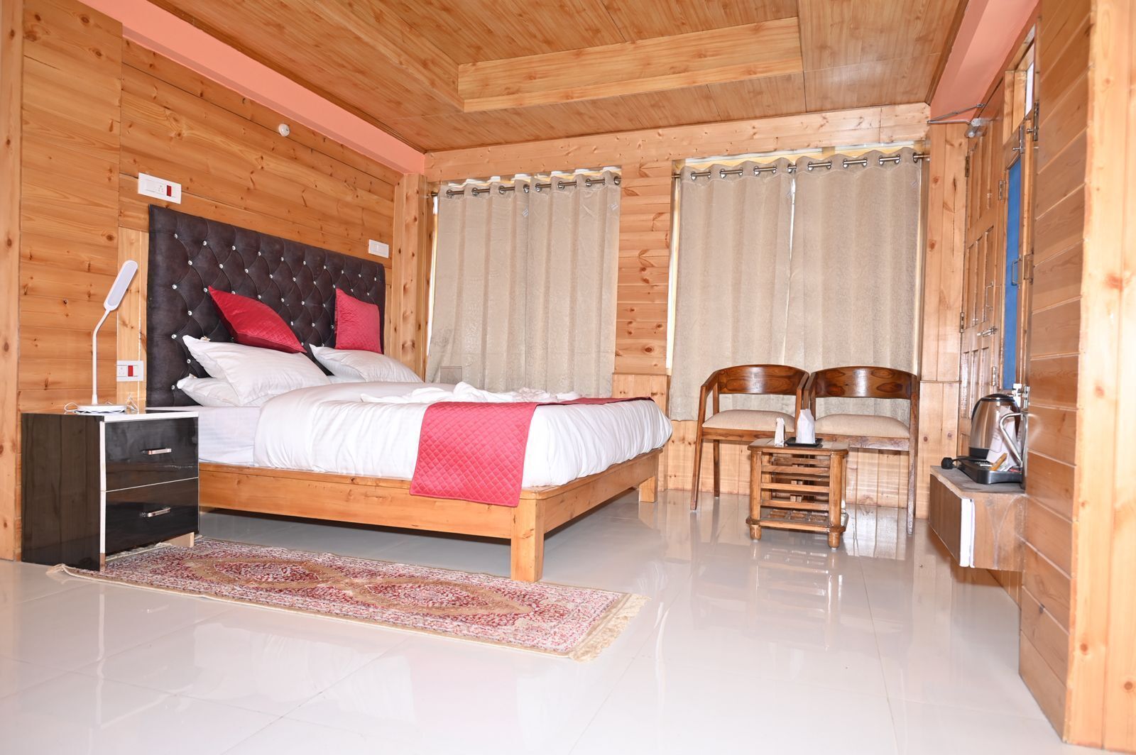 A wooden-paneled bedroom with a bed, two chairs, a nightstand, and a small table with a kettle.