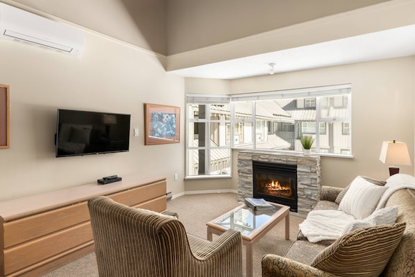 A living room with a flat-screen TV, a fireplace, a sofa, a coffee table, and a window with a view of buildings.