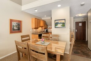 A dining area with a wooden table and chairs, a vase with flowers, and a kitchen in the background. - Glacier Lodge 340 NEW central AC just installed 2026 (Whistler)