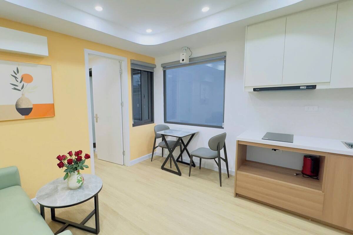 Un espacio habitable compacto con sofá, una mesita redonda con un florero, comedor con dos sillas, zona de cocina con mostrador y alacenas, y un ventanal grande.