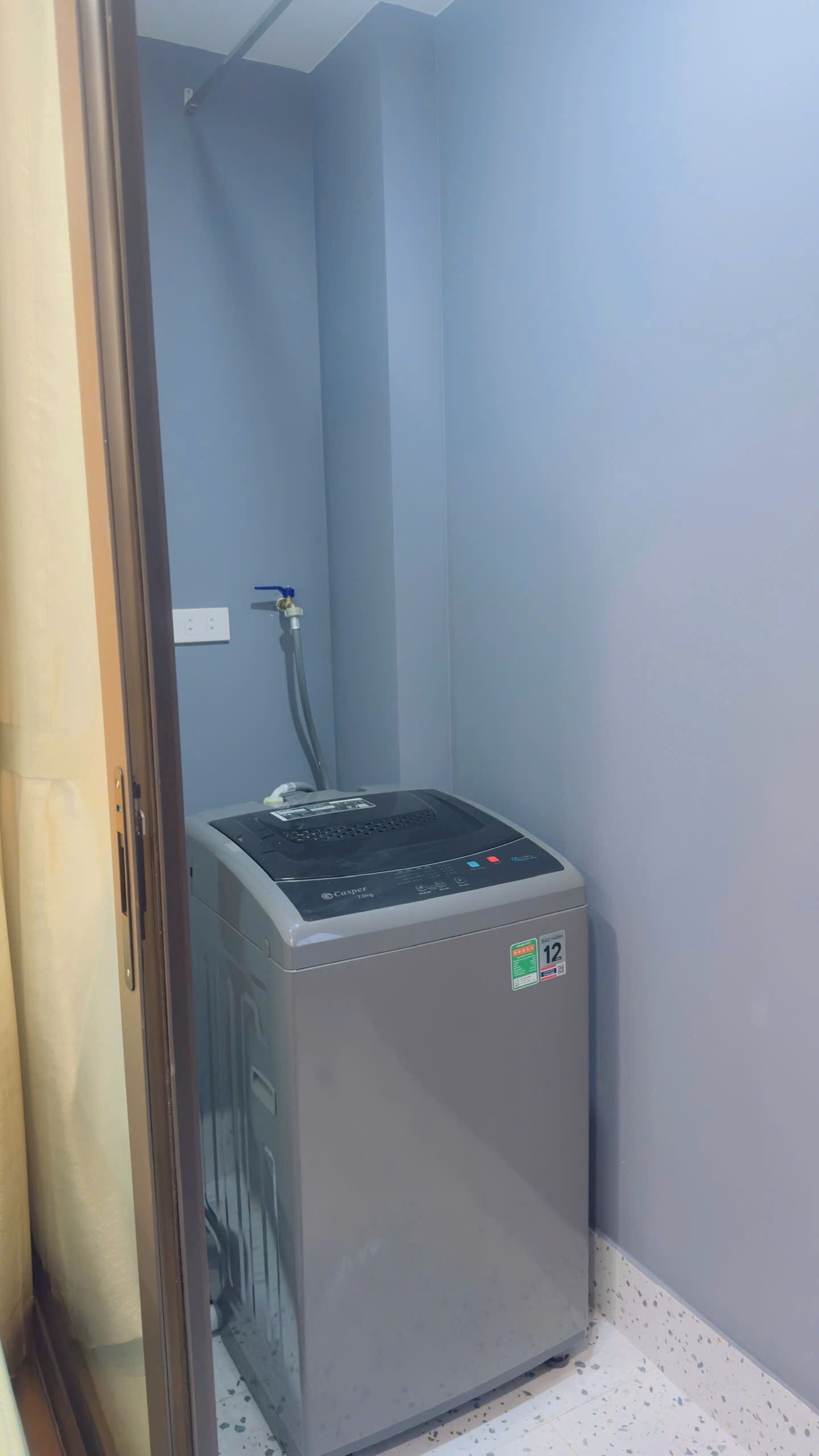 Calentador de agua compacto y gris en un rincón de una habitación con paredes azul claro y piso de baldosas blancas.
