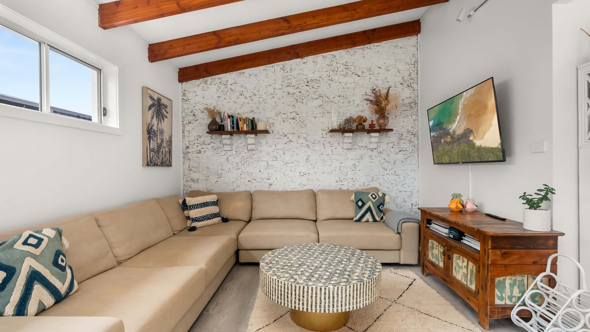 A living room with a beige sectional sofa, a round coffee table, a wooden cabinet, and a white brick wall.