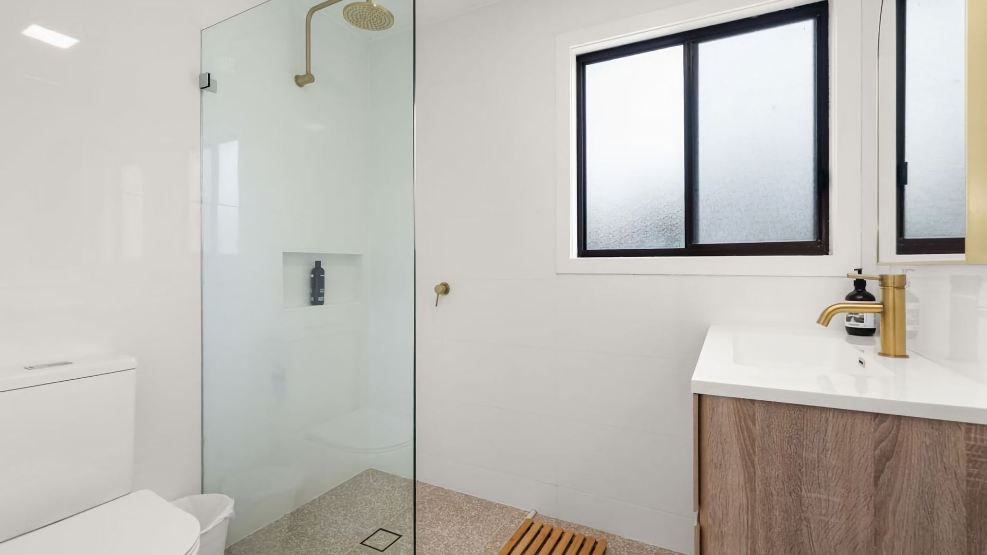 A modern bathroom with a glass shower enclosure, a white sink with a gold faucet, and a window above the mirror.