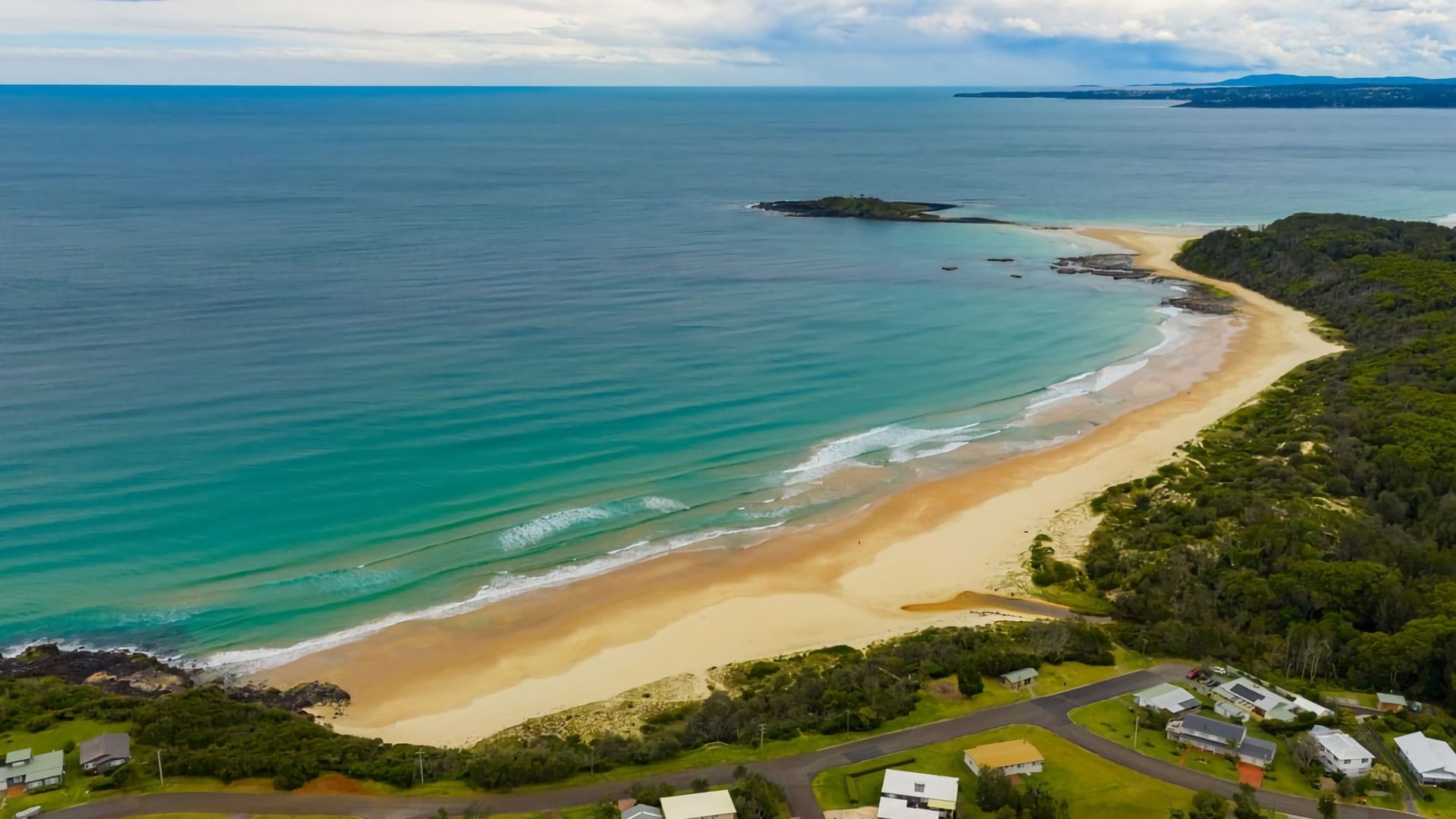 Vue aérienne d’une zone côtière avec une plage de sable, un petit village composé de maisons, et une vue dégagée sur l’océan.
