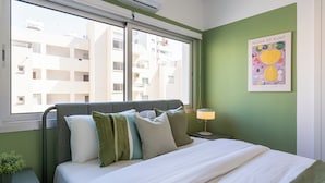 A hotel room with a bed, a nightstand, a lamp, a window with a view of buildings, and a poster on the wall. - Hazel 2-BR Apt. & Balcony in Larnaca (Larnaca)